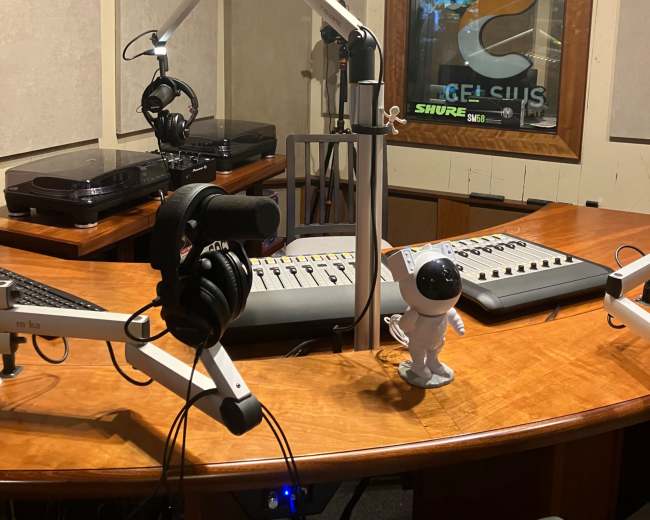 The image shows a radio studio with a wooden desk, audio equipment, microphones, headphones, and a small astronaut figurine.