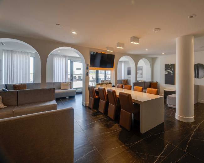 The image shows a modern interior space with a dining area featuring a large white table and brown chairs, surrounded by soft seating areas and large windows.