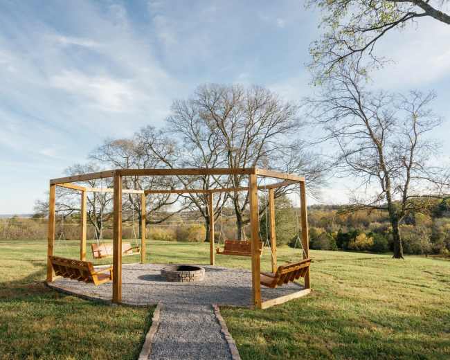 The image shows a wooden gazebo with swing benches and a stone fire pit, situated in an open grassy area surrounded by trees.