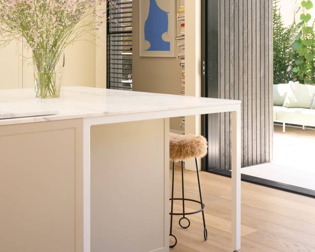 A modern kitchen features a marble island with a vase of flowers and an open door leading to an outdoor space filled with greenery.