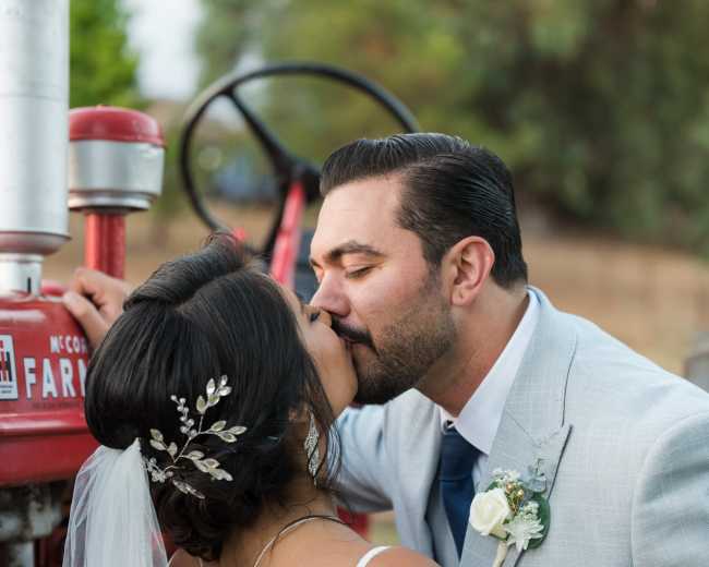 A couple shares a kiss while standing next to a vintage red tractor.