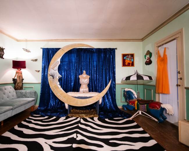 The image shows a whimsical room with a crescent moon backdrop, a mannequin in front of it, a patterned rug, and various decorations on the walls.