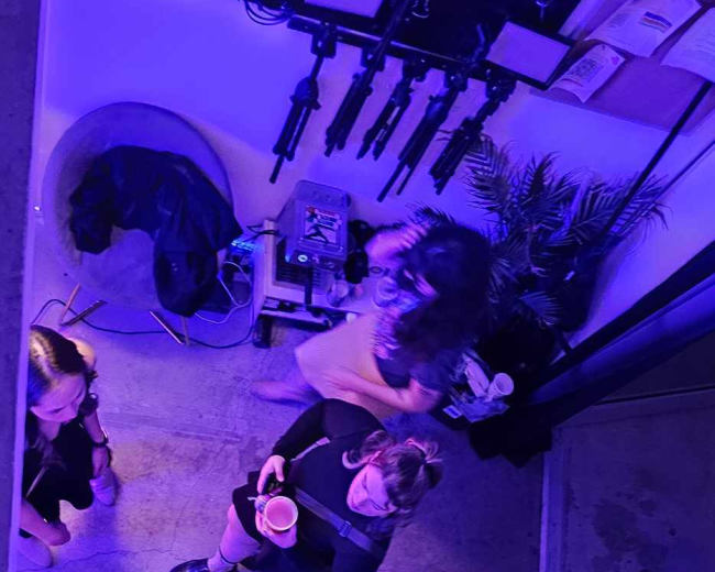 A group of people gathers in a dimly lit room filled with plants and colorful lighting, engaged in conversation near a sofa and various electronic equipment.