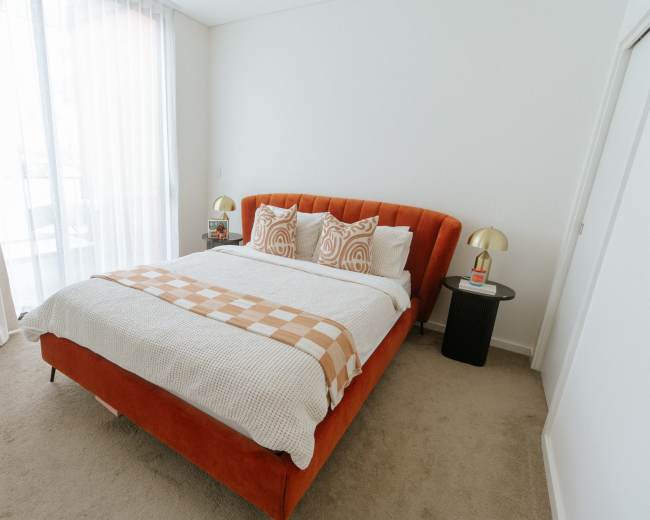 The bedroom features an orange upholstered bed with layered bedding, two patterned pillows, and a bedside table with a lamp.