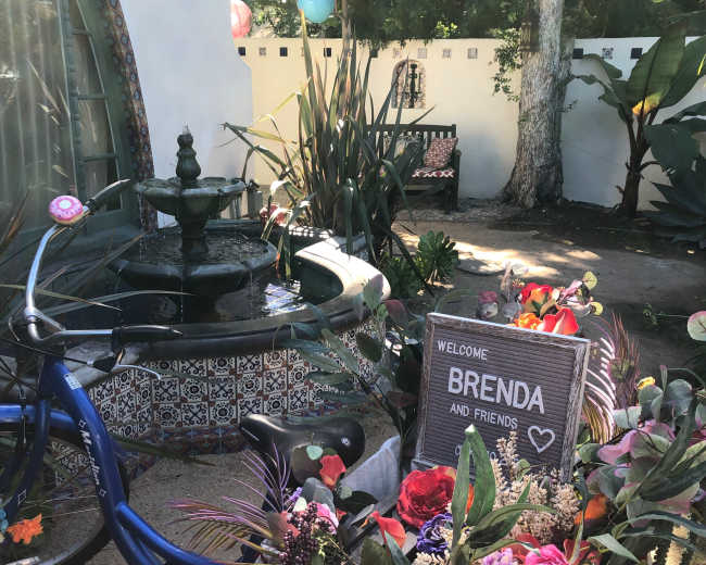 A blue bicycle decorated with flowers is leaning against a tiled fountain surrounded by greenery and a welcoming sign that reads "WELCOME BRENDA AND FRIENDS."