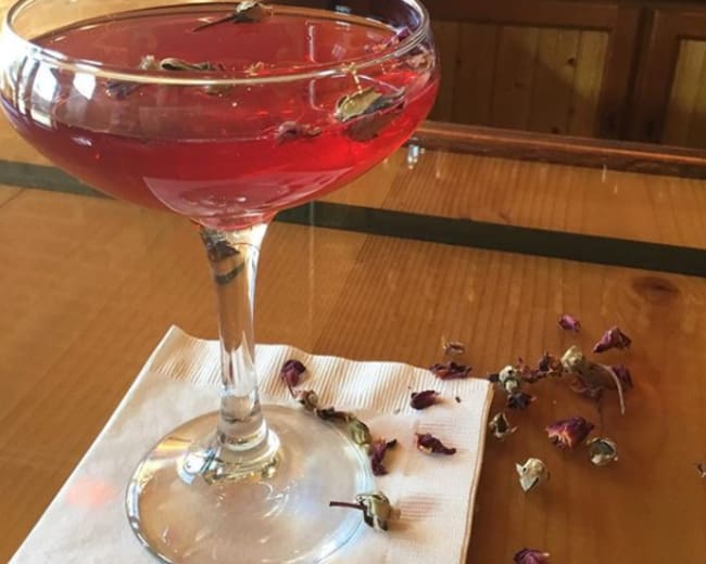 A cocktail glass filled with a red drink and garnished with dried rose petals rests on a wooden bar top beside scattered flower petals.
