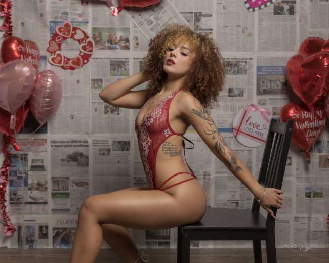 A person poses on a chair against a backdrop of newspaper clippings and Valentine's Day decorations, including heart-shaped balloons.