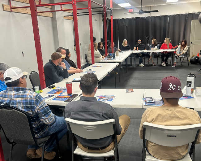 A group of people is seated around tables in a room set up for a meeting, with red exercise equipment in the background.