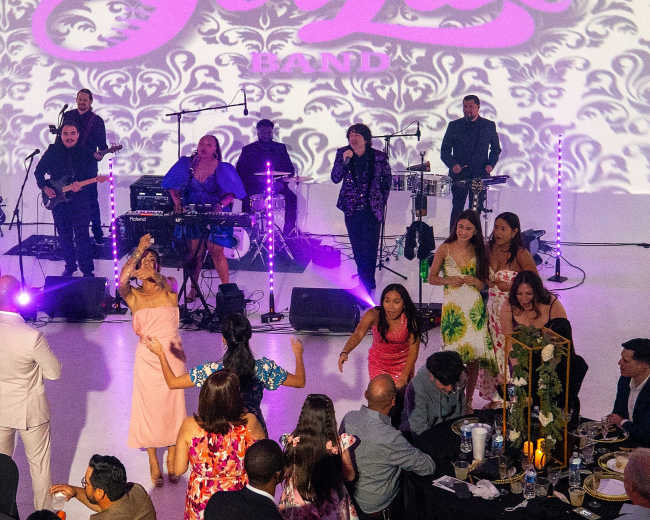 A lively band performs on stage while guests enjoy dinner and dance at a festive event.