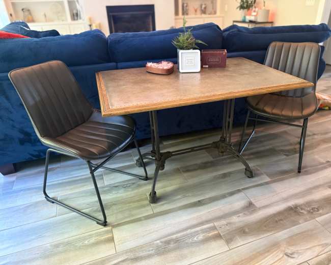 A rectangular dining table with a wooden top and metal legs is set between two leather chairs in a room with wooden flooring and a blue couch in the background.