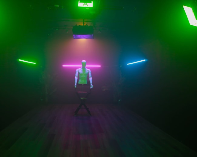 A mannequin stands in a dimly lit studio, illuminated by colorful neon lights and positioned on a stool.