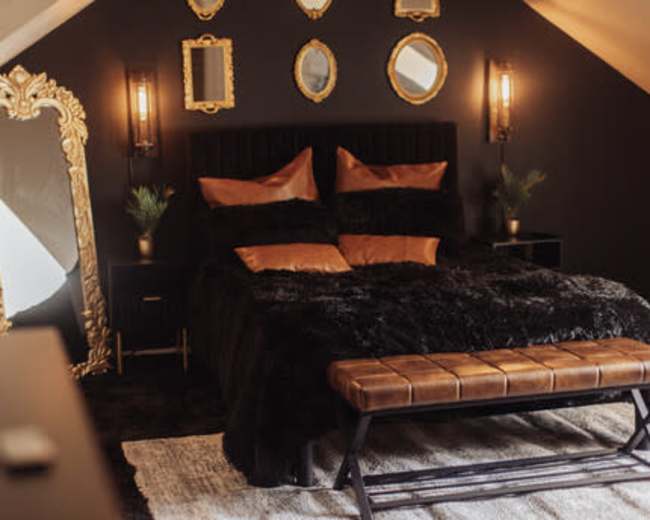 The image shows a stylish bedroom with a large bed covered in a black fur blanket, flanked by decorative mirrors on the walls and a bench at the foot of the bed.