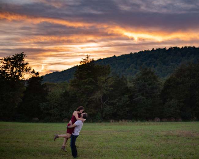 A couple embraces in a field at sunset, surrounded by trees and rolling hills.