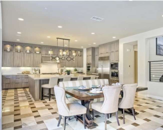 The image shows a modern kitchen and dining area, featuring a large dining table surrounded by upholstered chairs, with a staircase in the background and tiled flooring.