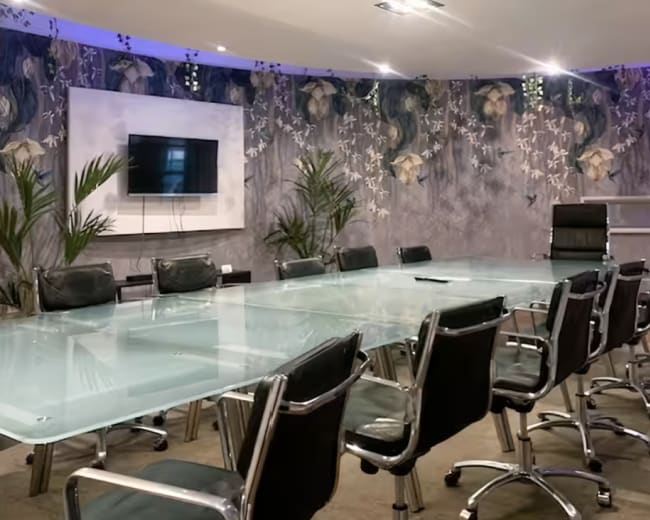 A modern conference room features a large glass table surrounded by black ergonomic chairs, with floral-patterned walls and a mounted screen for presentations.