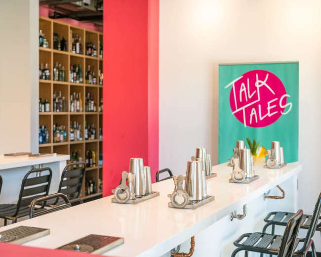 The image shows a sleek bar area with a white countertop, metal shaker sets lined up, and a colorful backdrop featuring shelves filled with various bottles.