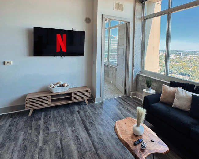 The living room features a television displaying the Netflix logo, a sofa with decorative pillows, a coffee table, and a large window offering a view of the skyline.