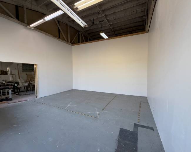 The image shows an empty room with white walls and a concrete floor, featuring a doorway on one side and overhead lighting fixtures.