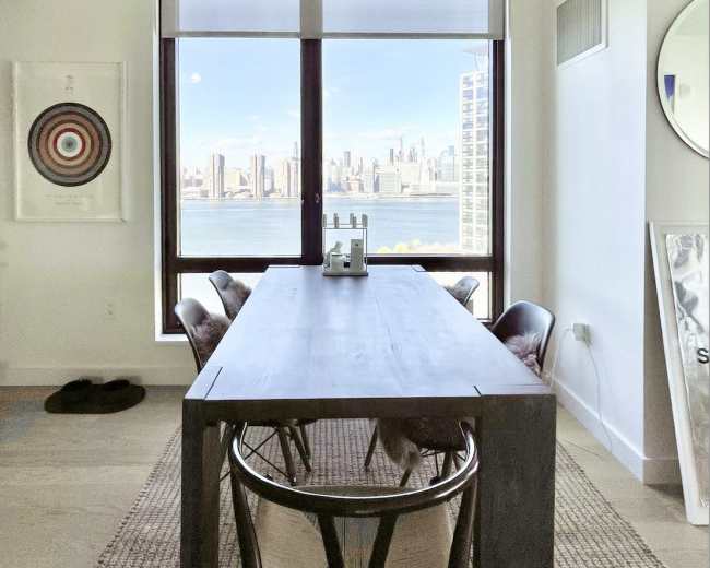 A long wooden dining table is set in a bright room with large windows showcasing a city skyline view.