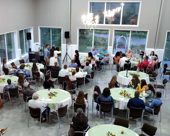 A gathering is taking place indoors in a spacious room with tables set for guests, while people engage in conversations and a few are presenting at the front.