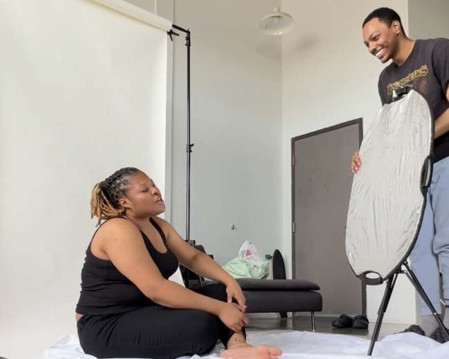 A woman sits on a white backdrop while a man sets up an ironing board in a well-lit room.