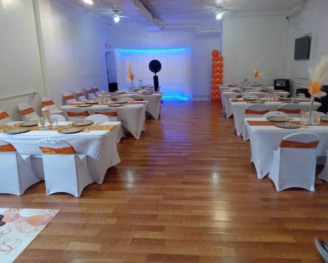 The image shows a banquet hall set up with multiple tables covered in white tablecloths and adorned with orange accents, organized for an event.