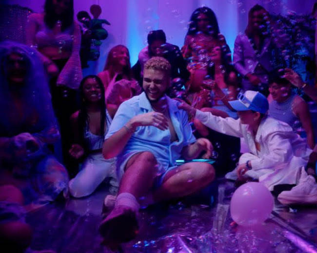 A group of people in colorful attire celebrate at a lively party, surrounded by bubbles and balloons, with one person sitting prominently on the floor.