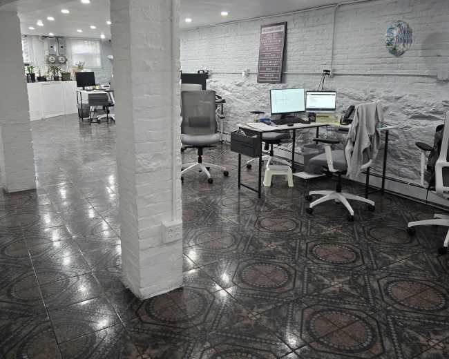 The image shows a well-lit office space featuring multiple desks with computers, ergonomic chairs, and a patterned tiled floor.