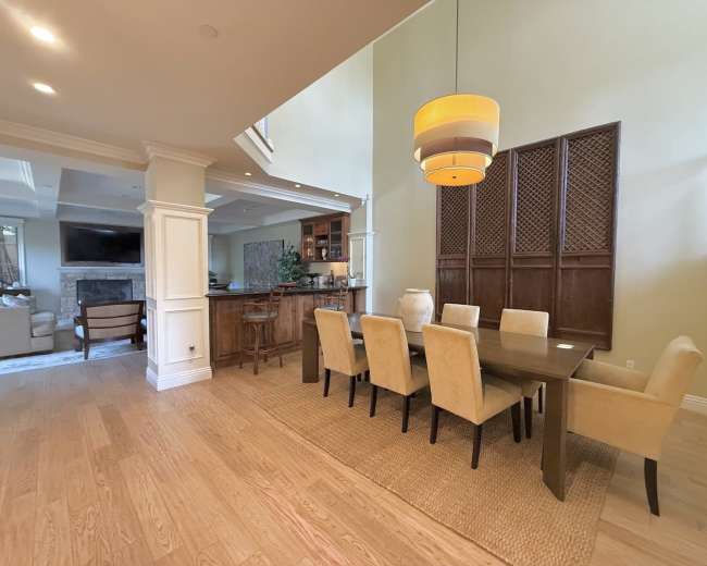 The image shows a spacious dining area featuring a long wooden table surrounded by upholstered chairs, with a large light fixture hanging above.