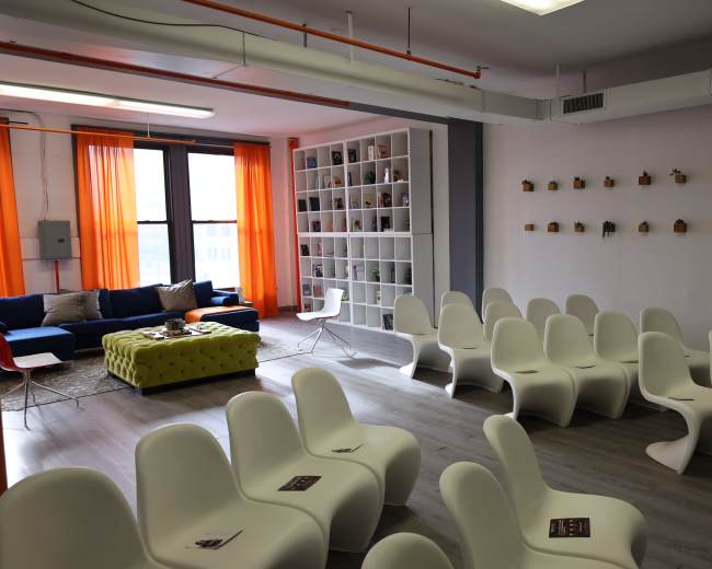 The image shows a modern room with white chairs arranged in rows, a colorful couch, a coffee table, and decorative shelves on the walls.