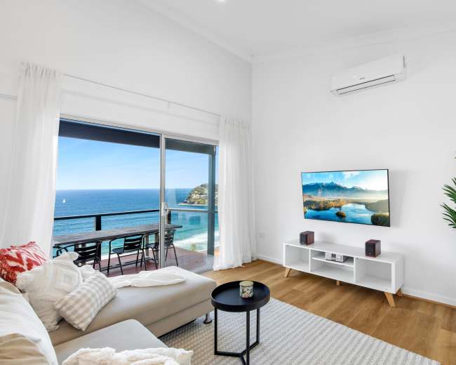 The image shows a modern living room with a large window offering a view of the ocean, featuring a sofa, a coffee table, and a television on a white cabinet.