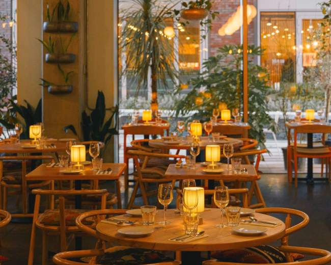The image shows a warmly lit restaurant interior with wooden tables set for dining, surrounded by plants and large windows.