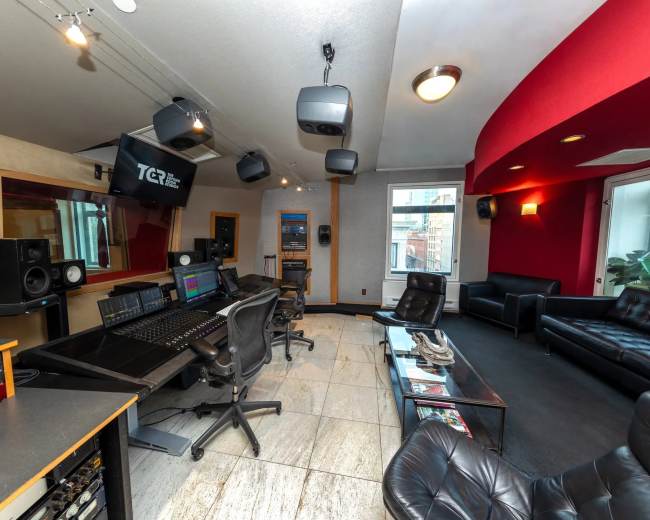 The image shows a modern recording studio featuring a mixing console, multiple speakers, and seating areas.