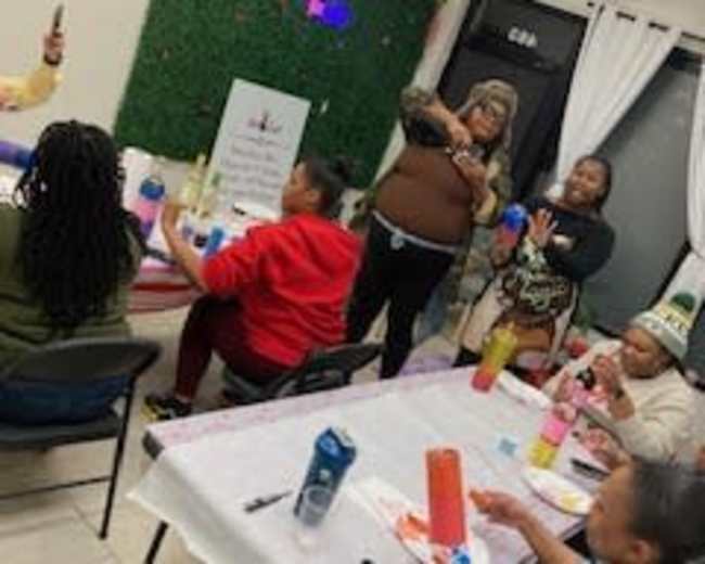 A group of people are engaged in a craft activity around tables in a brightly lit room decorated with a green wall display.