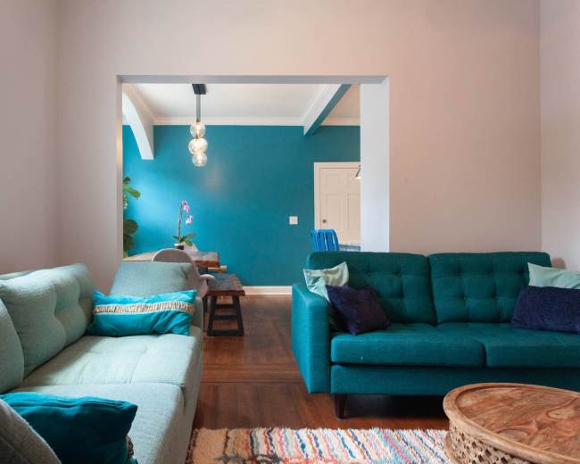A living room features two teal sofas, a wooden coffee table, and a blue accent wall, with a doorway leading to a dining area.