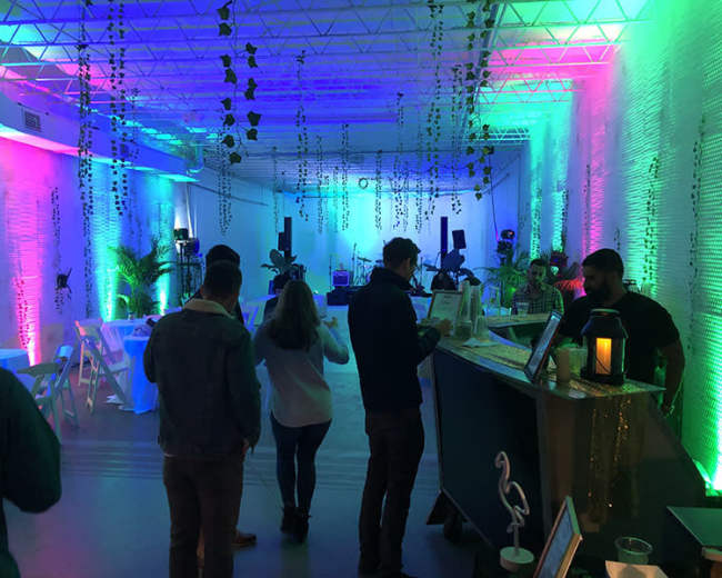 The scene depicts a brightly lit event space with colorful lighting, tables arranged for guests, and people mingling in the foreground.