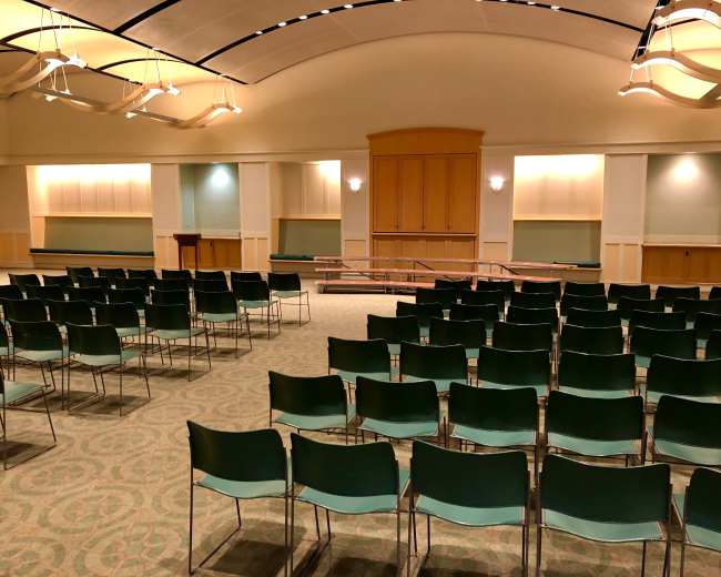 The image shows an empty indoor auditorium with rows of green chairs facing a wooden stage at the front.