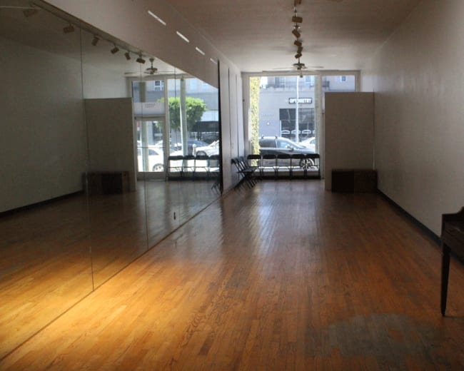 The image shows a spacious room with wooden flooring, large mirrors on one side, and natural light coming in through a window.