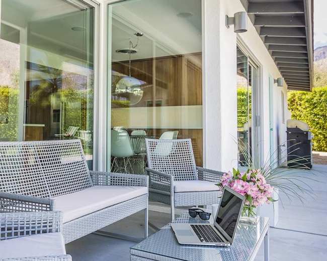 A modern patio features wicker furniture, a laptop on a small table, and a flower arrangement, with a view of a green hedge and glass doors.