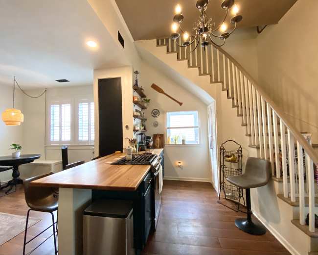 A modern kitchen features a wooden countertop, stainless steel appliances, and an adjacent dining area with a staircase and decorative chandelier.