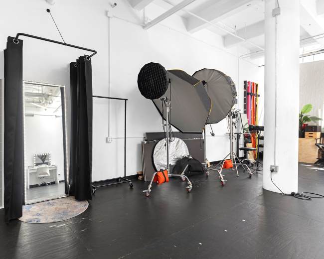 The image shows a spacious studio interior with a large mirror, photography lighting equipment, and a minimalist black floor.