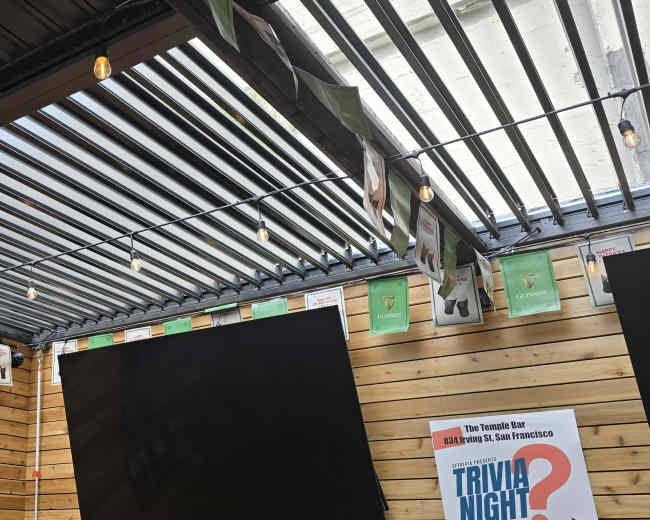 The image shows a sports bar interior with a large screen displaying a basketball game, surrounded by wooden walls and a skylight featuring string lights.