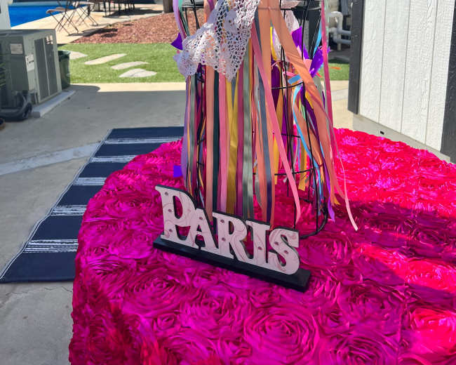A decorative mannequin draped with colorful ribbons and flowers stands on a table covered in bright pink fabric, with a sign reading "PARIS" in front of it.