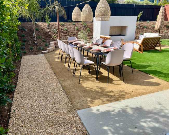 A patio area with a long dining table set for a meal, surrounded by green grass and decorative plants.