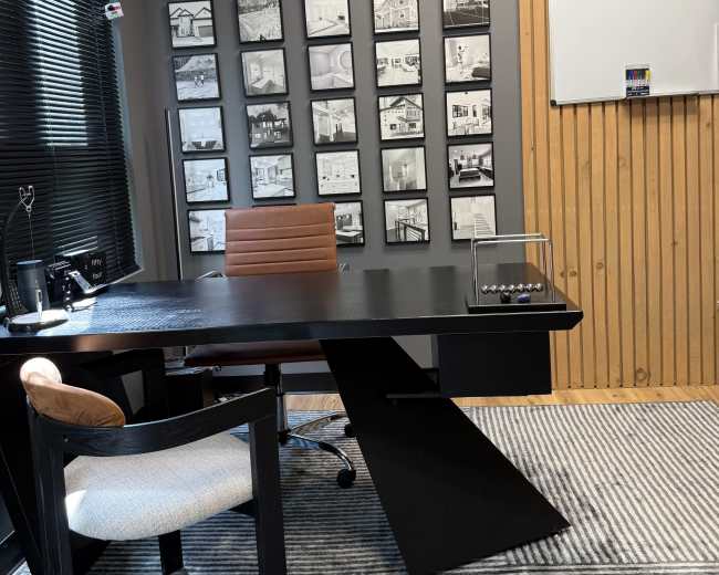 The image shows a modern office with a black desk, a brown chair, and a wall decorated with multiple black-and-white framed photographs.