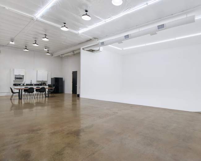 The image shows a spacious studio with a white backdrop, polished concrete floor, and a kitchenette area with black chairs and cabinets in the corner.