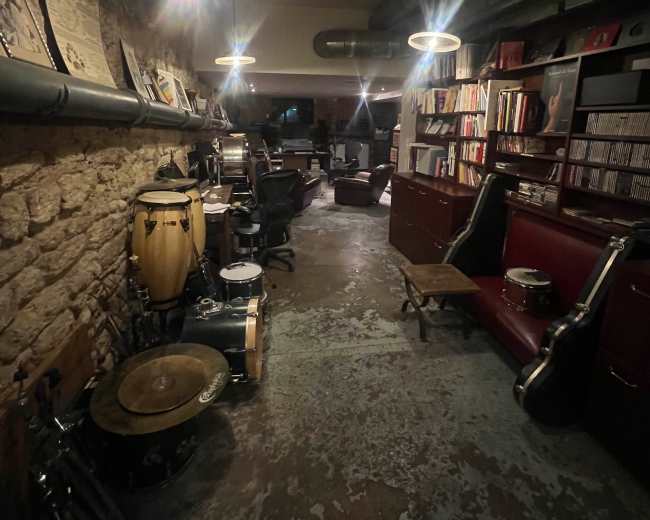 The image shows a dimly lit music studio with stone walls, various musical instruments, and shelves filled with records and books.