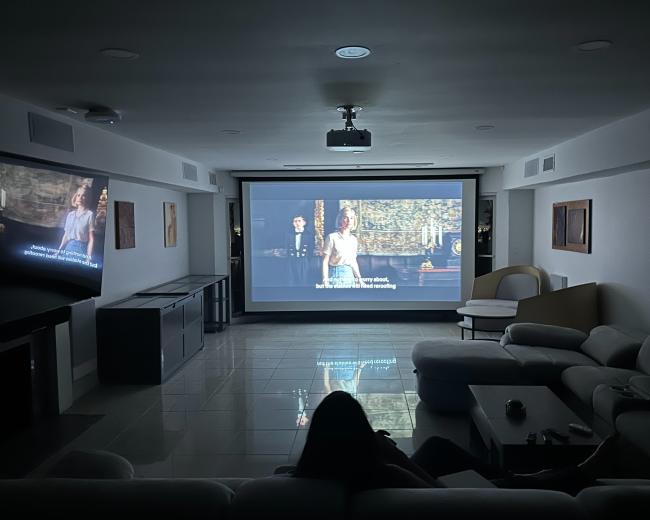 A dimly lit room features two projection screens displaying a movie, with a person seated on a white sofa in front of them.