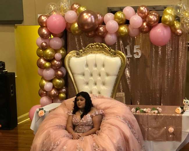 A girl in a pink gown sits on an ornate, white throne in front of a decorated backdrop with balloons.