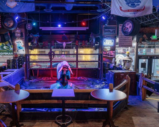 The image shows a dimly lit wrestling ring surrounded by wooden tables, decorated with various flags and neon lights, inside a lively venue.